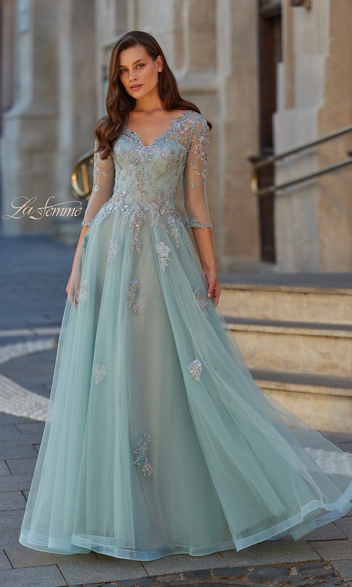 A woman stands outdoors on a paved walkway wearing the La Femme 33536 long formal dress in dusty blue, featuring tulle fabric, floral embroidery, and sheer sleeves.
